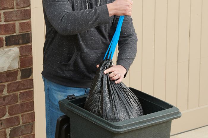 Person pulling the drawstring on an ultra strong large trash bag while placing it in a large trash can.